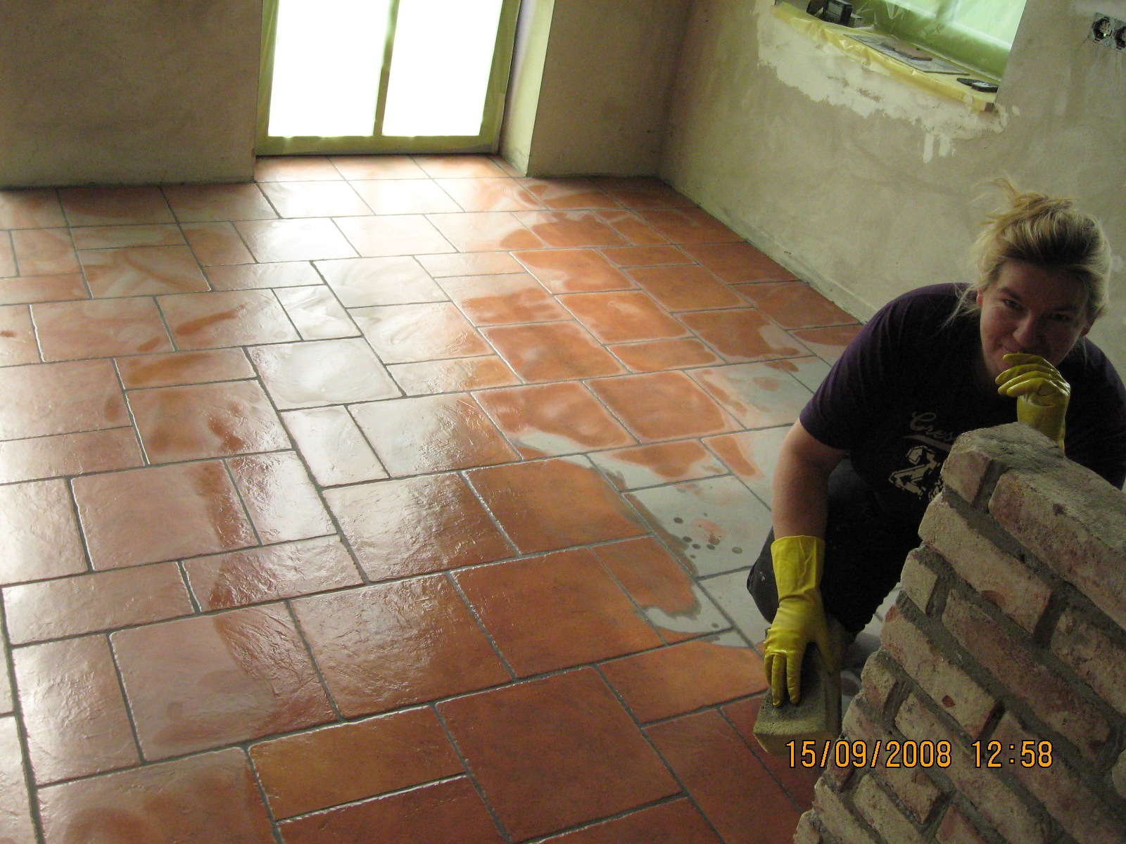 Kobieta w żółtych rękawicach czyści gąbką nowo położoną, brązową terakotę na podłodze pomieszczenia podczas prac wykończeniowych, widoczne resztki fugi, ściany przygotowane do malowania, okno...