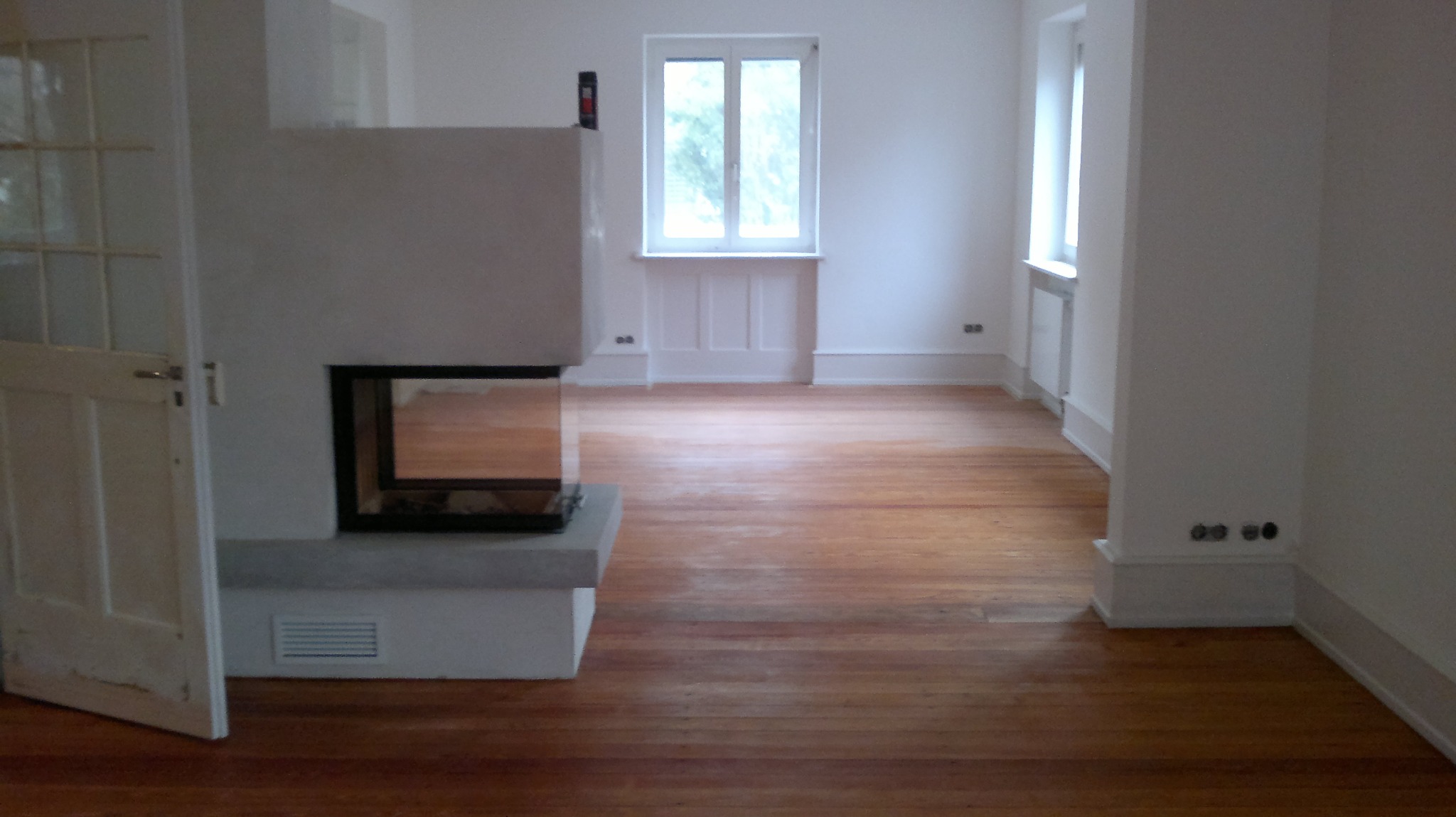 Pusty pokój z drewnianą podłogą po renowacji, widoczny kominek z wkładem, białe ściany i stolarka okienna, częściowo otwarta biała drewniana drzwi po lewej stronie.