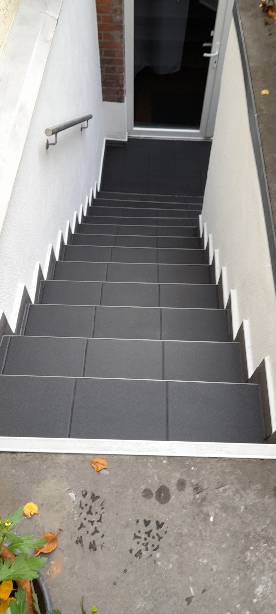 Schody wyłożone ciemnymi płytkami z białymi listwami wykończeniowymi, prowadzące do drzwi wejściowych. Widoczna metalowa poręcz i fragment elewacji z cegły. Na posadzce ślady po deszczu i opadłe...