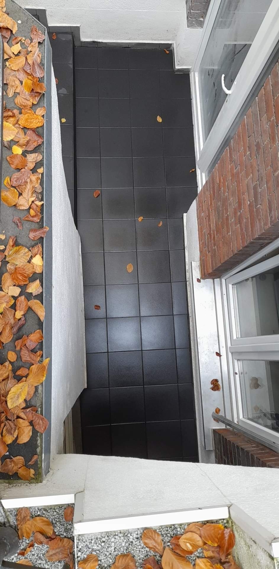 Ciemne, kwadratowe płytki na balkonie z opadłymi, brązowymi liśćmi jesiennymi, widok z góry, częściowo widoczne okno z ceglaną obudową.