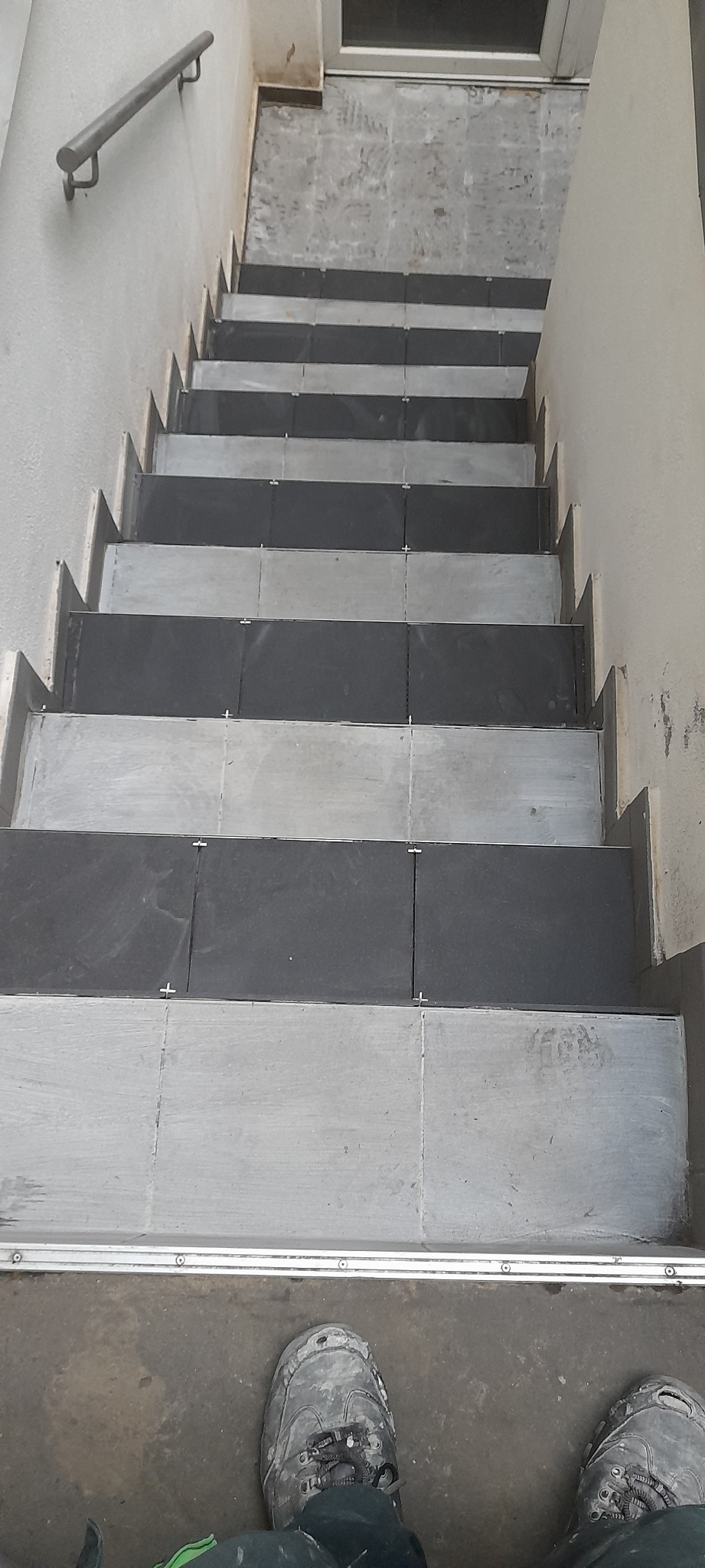 Wykończone schody z szaro-czarną glazurą, widok z góry, z fragmentem butów roboczych na dole kadru.