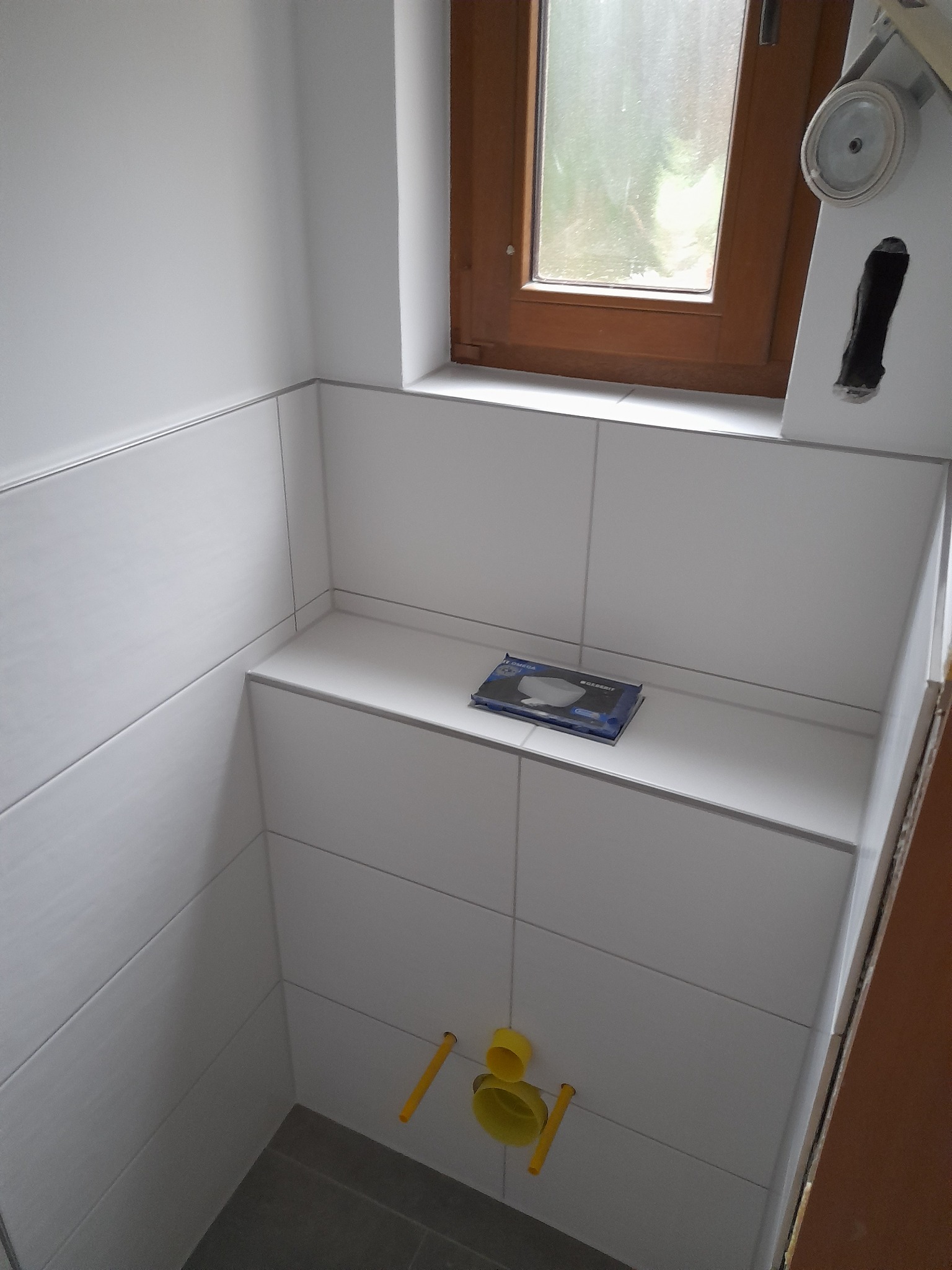 Wnętrze łazienki w trakcie wykańczania: białe płytki ścienne i podłogowe, okno z brązową ramą, żółte rury instalacyjne, pudełko z produktem Geberit na półce.