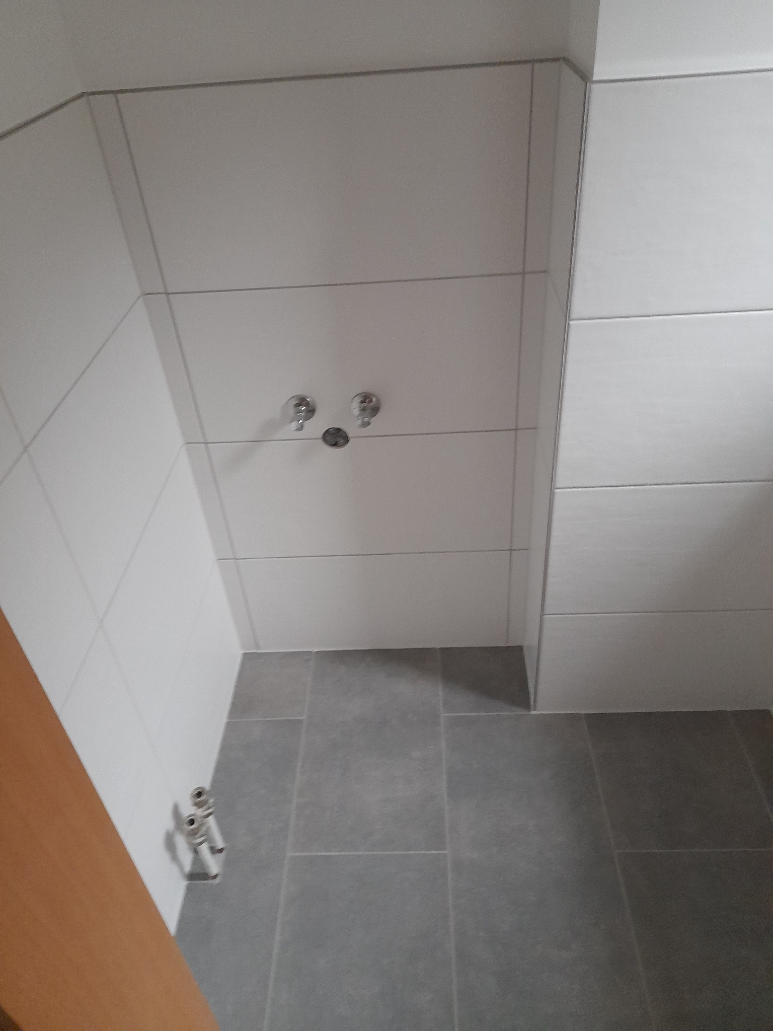 Wykończona łazienka z białymi, prostokątnymi płytkami na ścianach i szarymi, kwadratowymi płytkami na podłodze, widoczne rury doprowadzające wodę do baterii prysznicowej.