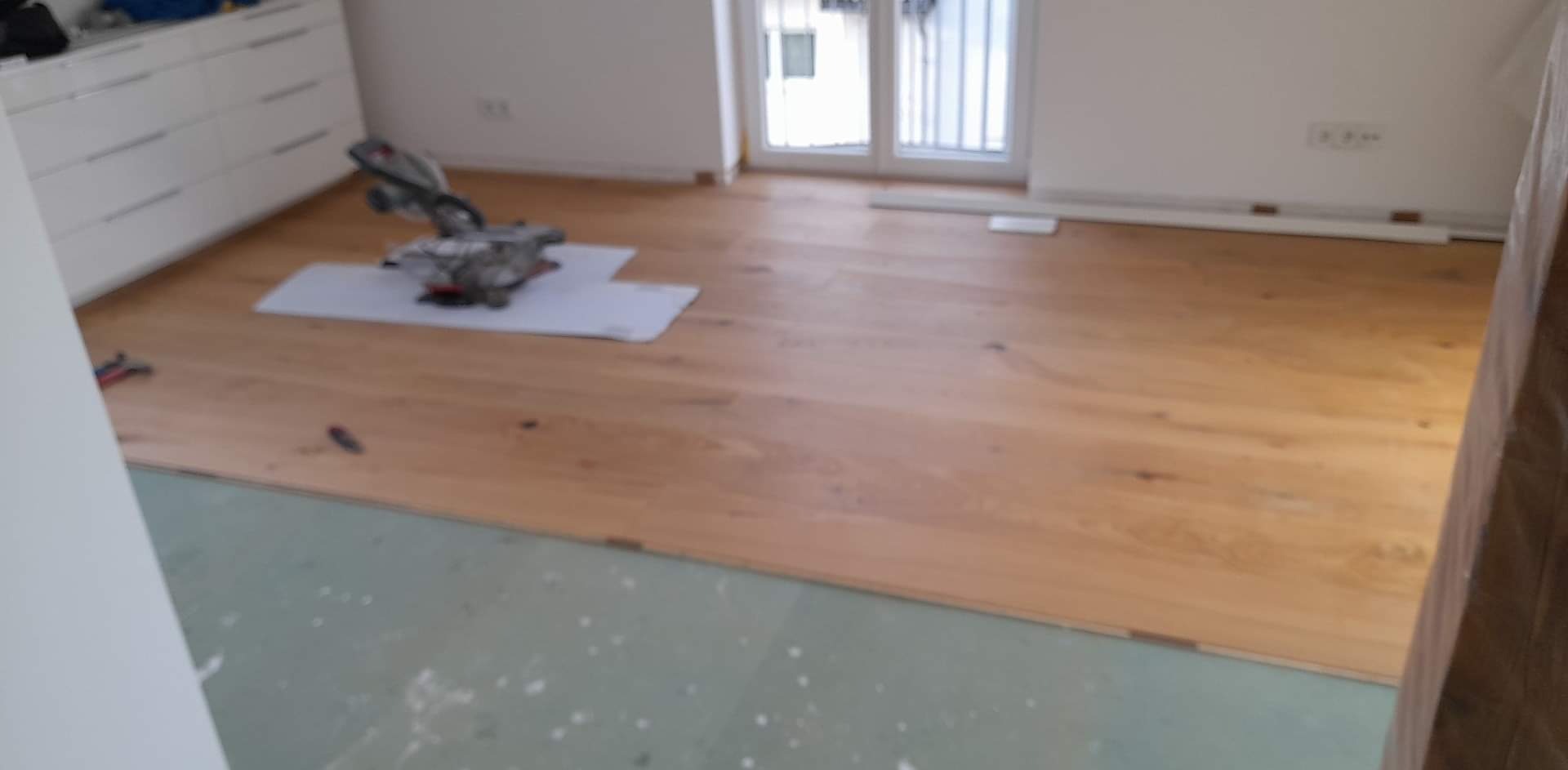 Proces układania jasnej, drewnianej podłogi w pokoju, widoczna piła do cięcia paneli na białym podkładzie.