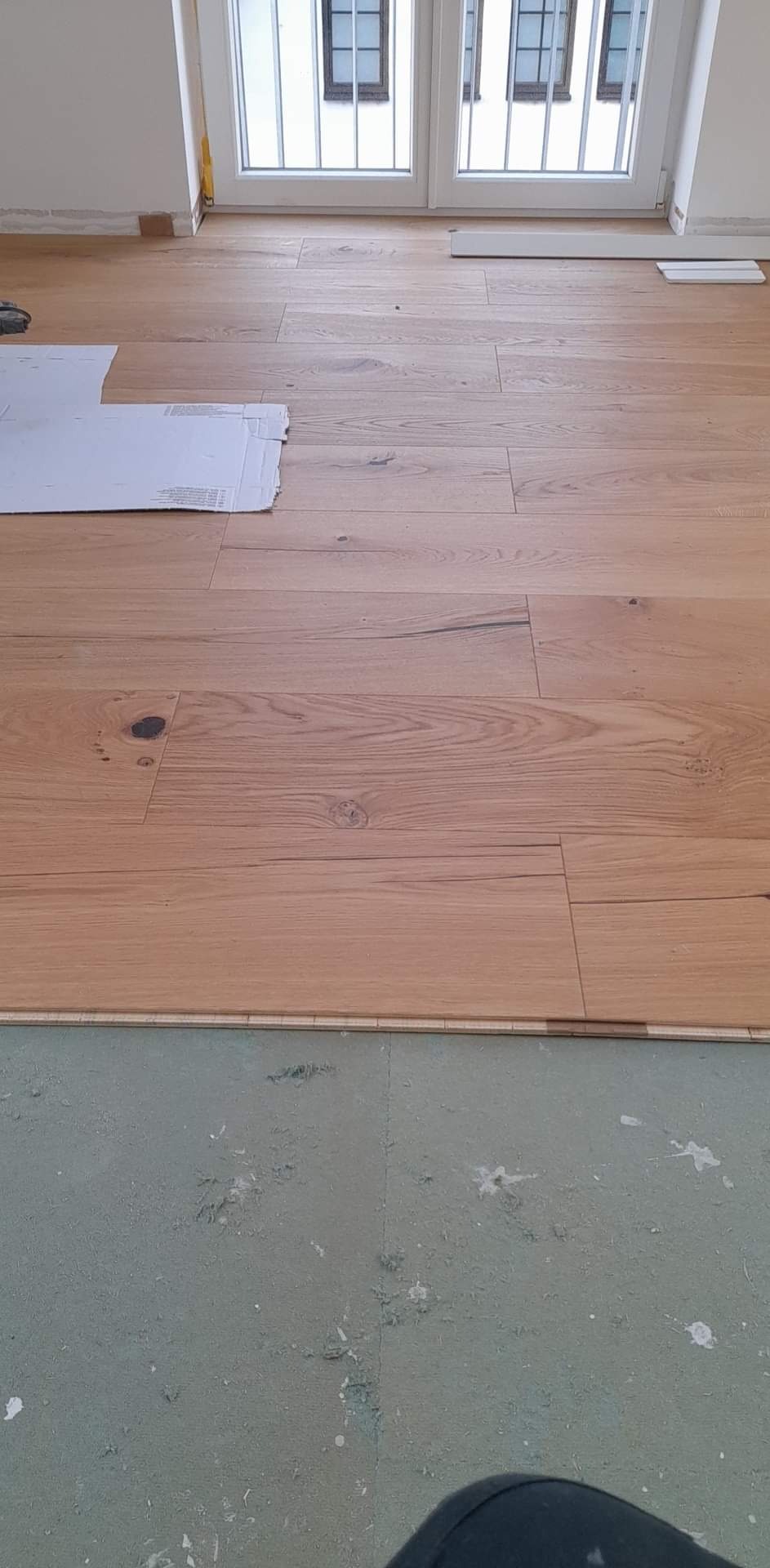 Układanie parkietu z jasnego drewna, widoczne deski w trakcie montażu, podkład pod parkiet i białe okno w tle.