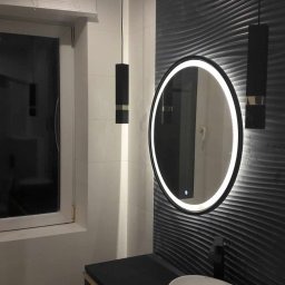 Nowoczesna łazienka: owalne lustro z podświetleniem LED na tle ściany z dekoracyjną fakturą, minimalistyczne lampy wiszące i fragment blatu z umywalką.
