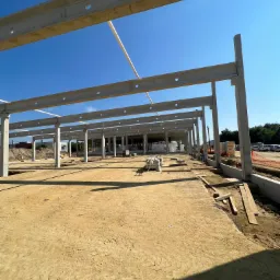 Konstrukcja szkieletowa budynku w trakcie budowy, widoczne betonowe słupy i belki stropowe na tle błękitnego nieba.