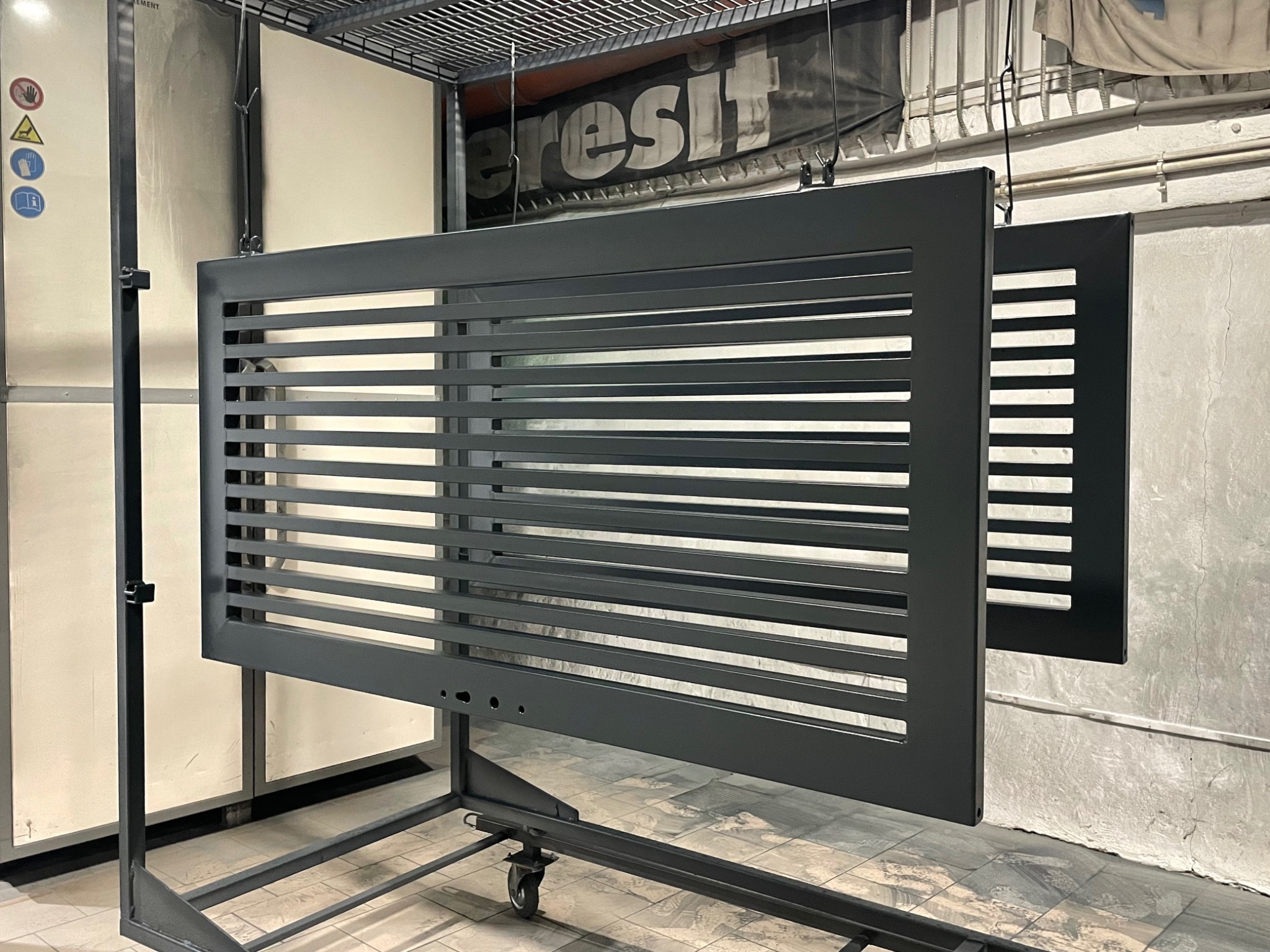 Dwie metalowe bramki, z poziomymi szczebelkami, zawieszone na hakach w hali produkcyjnej po nałożeniu matowej, szarej powłoki.