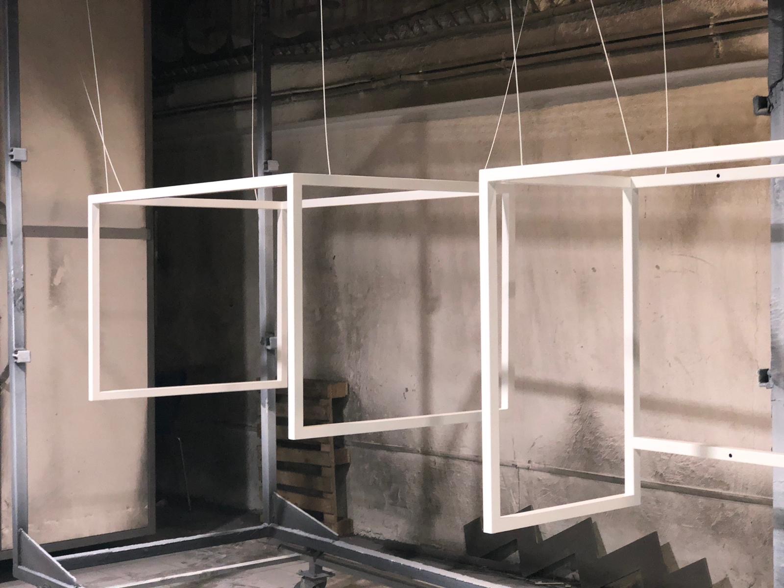 Białe, kwadratowe ramy zawieszone na linkach w hali, prawdopodobnie przed lub po malowaniu proszkowym, z widocznym wózkiem transportowym pod spodem.