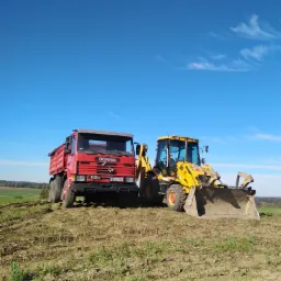 Czerwona ciężarówka Scania 113H i żółta koparko-ładowarka JCB na polu z widocznymi śladami opon i świeżą zielenią, błękitne niebo w tle.