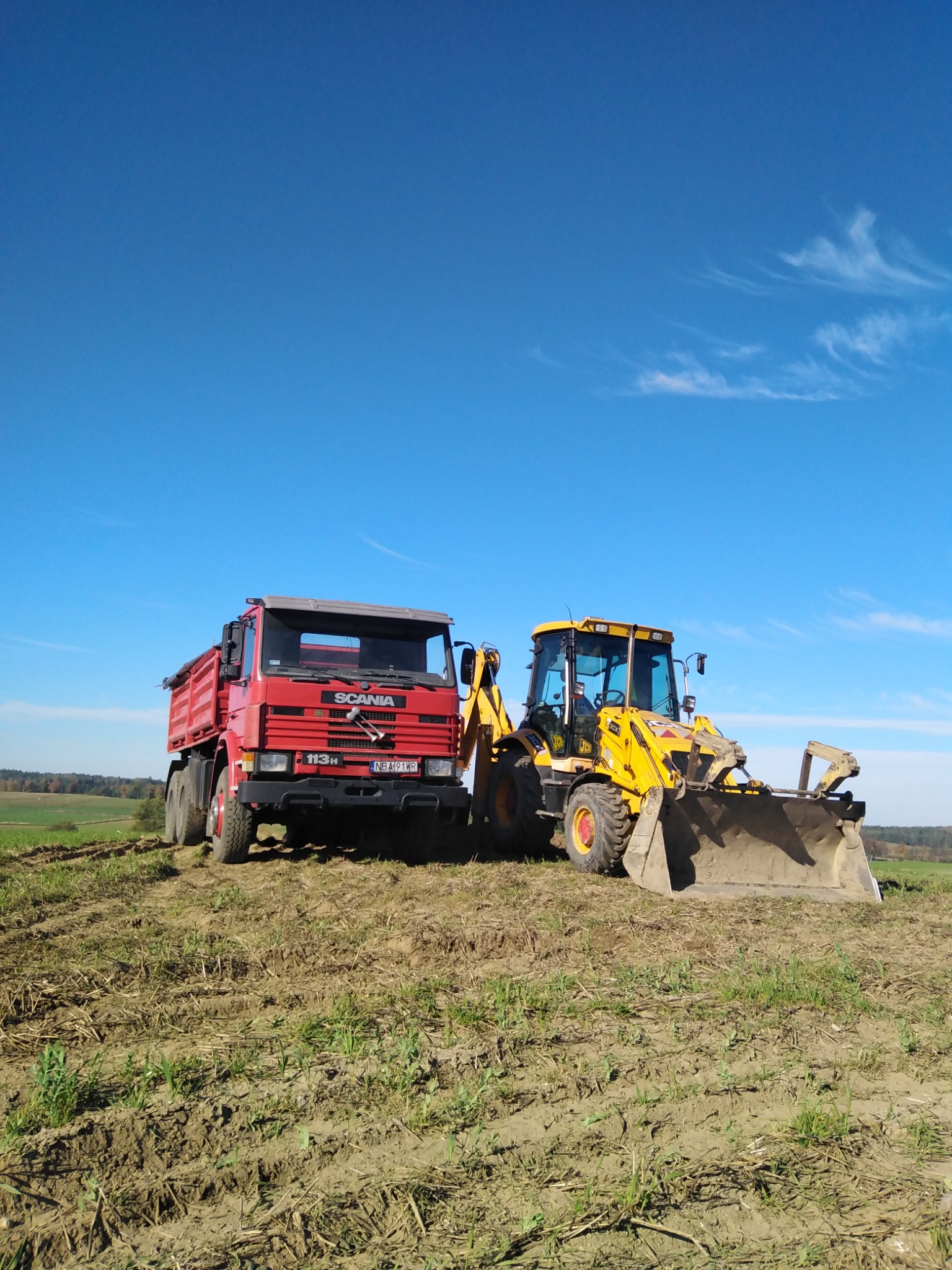 Czerwona ciężarówka Scania 113H i żółta koparko-ładowarka JCB na polu z widocznymi śladami opon i świeżą zielenią, błękitne niebo w tle.