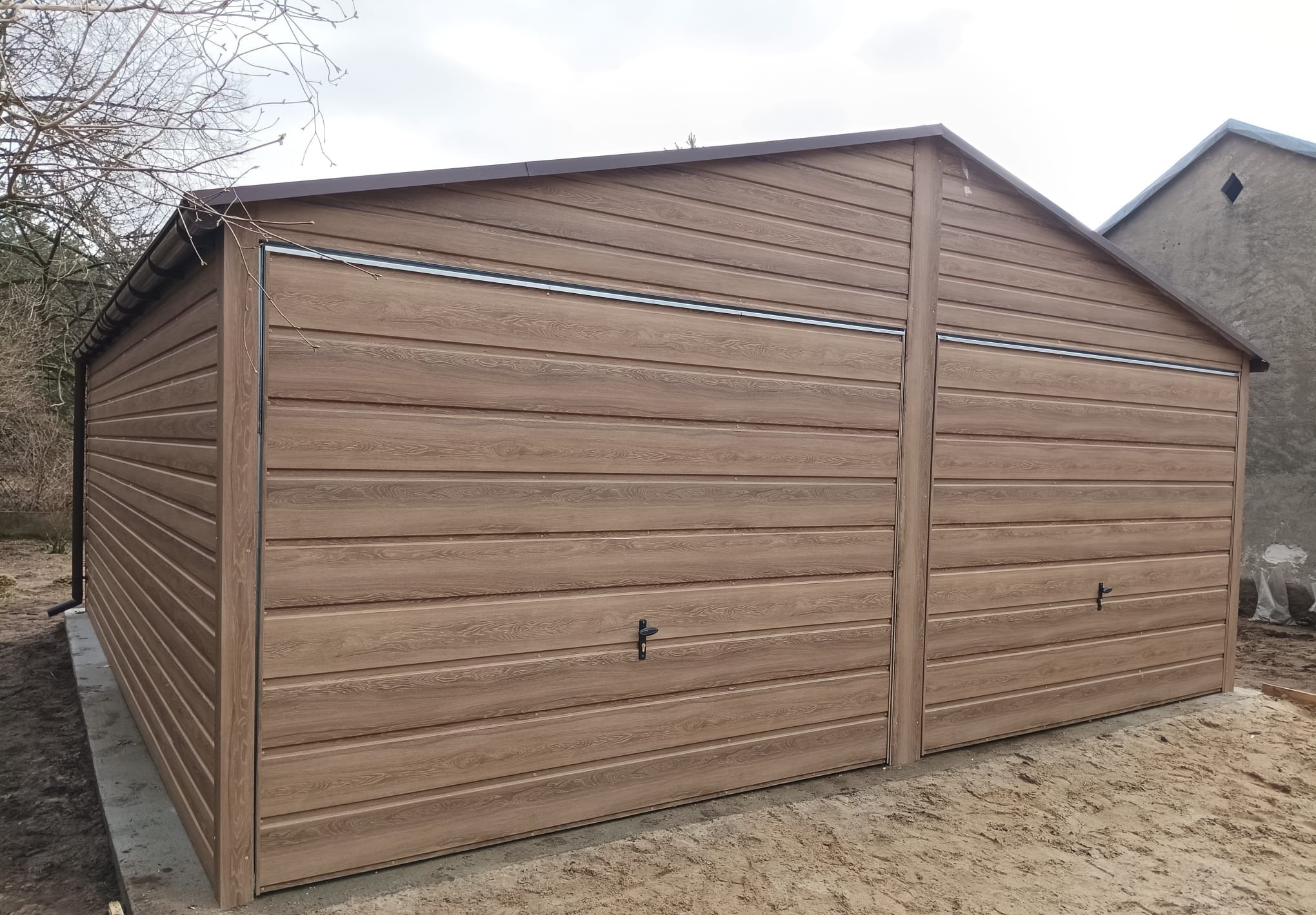 Dwustanowiskowy garaż z blachy imitującej drewno, z dwuspadowym dachem i dwoma parami drzwi uchylnych, na betonowej wylewce.