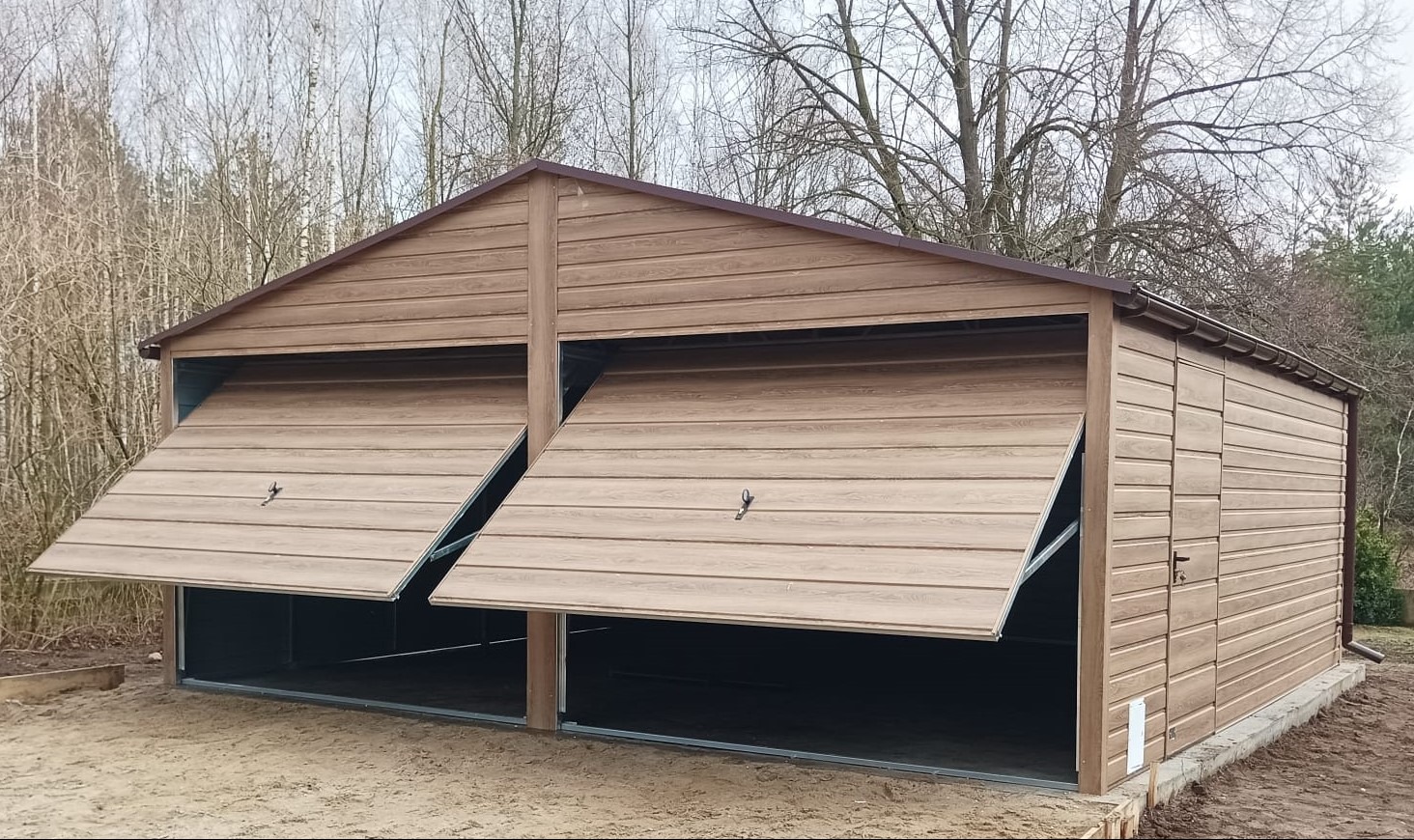 Dwustanowiskowy garaż z blachy imitującej drewno, z uchylonymi drzwiami, na tle drzew bezlistnych.