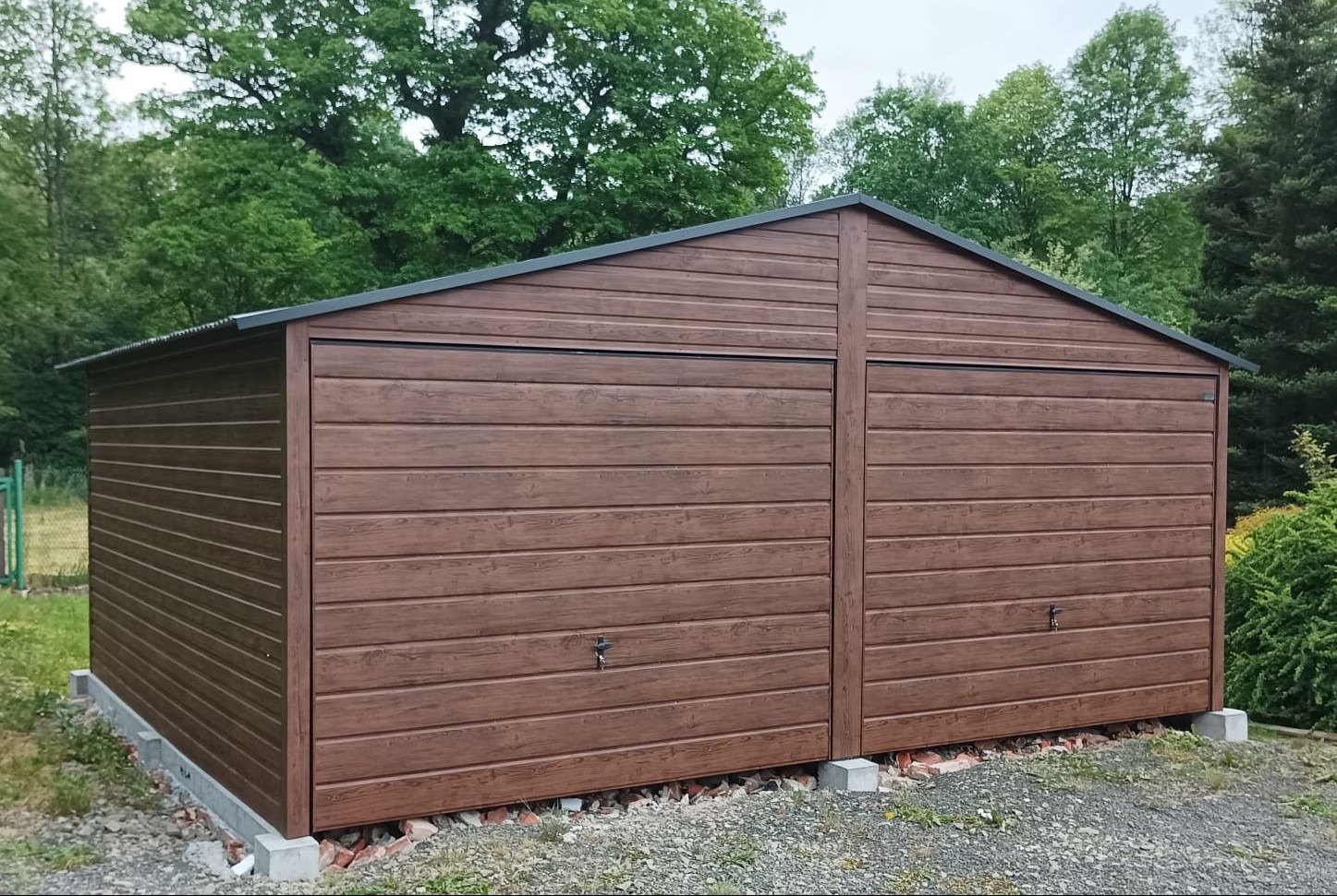 Dwustanowiskowy, brązowy garaż blaszany z dachem dwuspadowym w Szczyrzycu, z imitacją drewnianej elewacji, posadowiony na betonowych bloczkach.