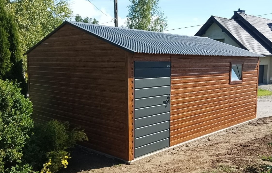 Brązowy garaż blaszany z szarą, poziomą bramą segmentową i małym oknem, w tle dom jednorodzinny.
