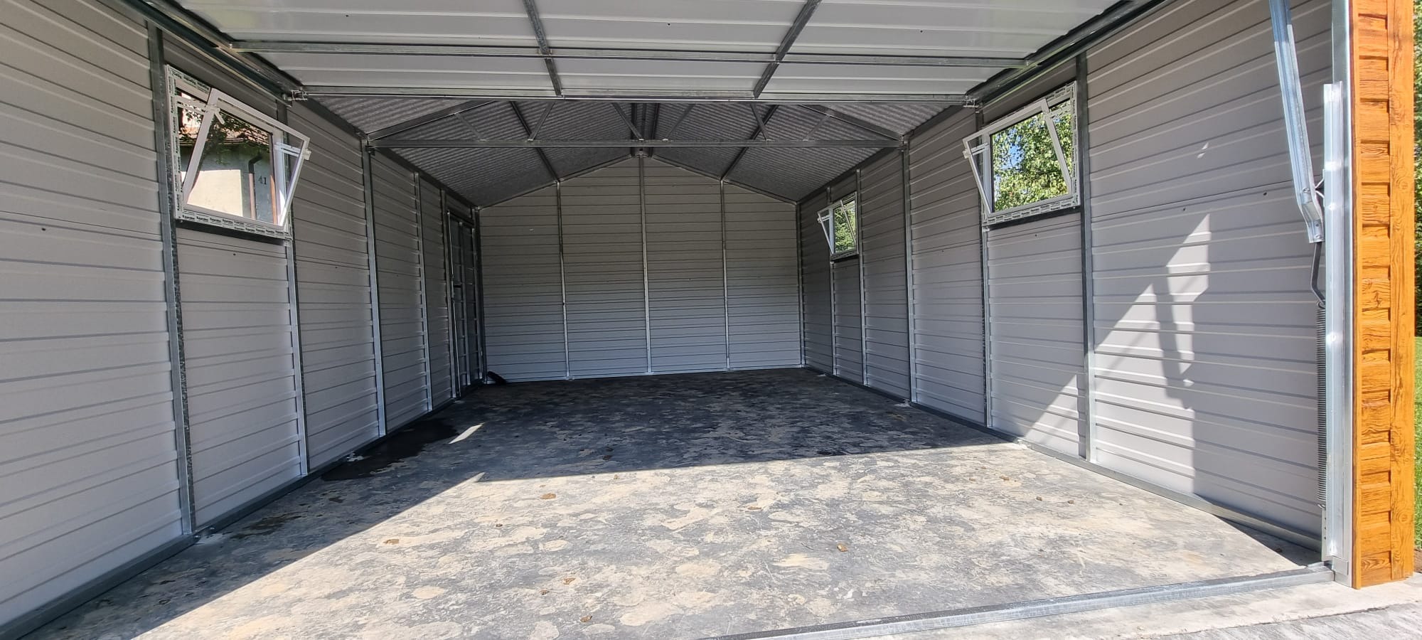 Wnętrze nowego, pustego garażu z blachy falistej, z dwoma uchylnymi oknami po bokach i betonową posadzką, widok z zewnątrz na jasne otoczenie.