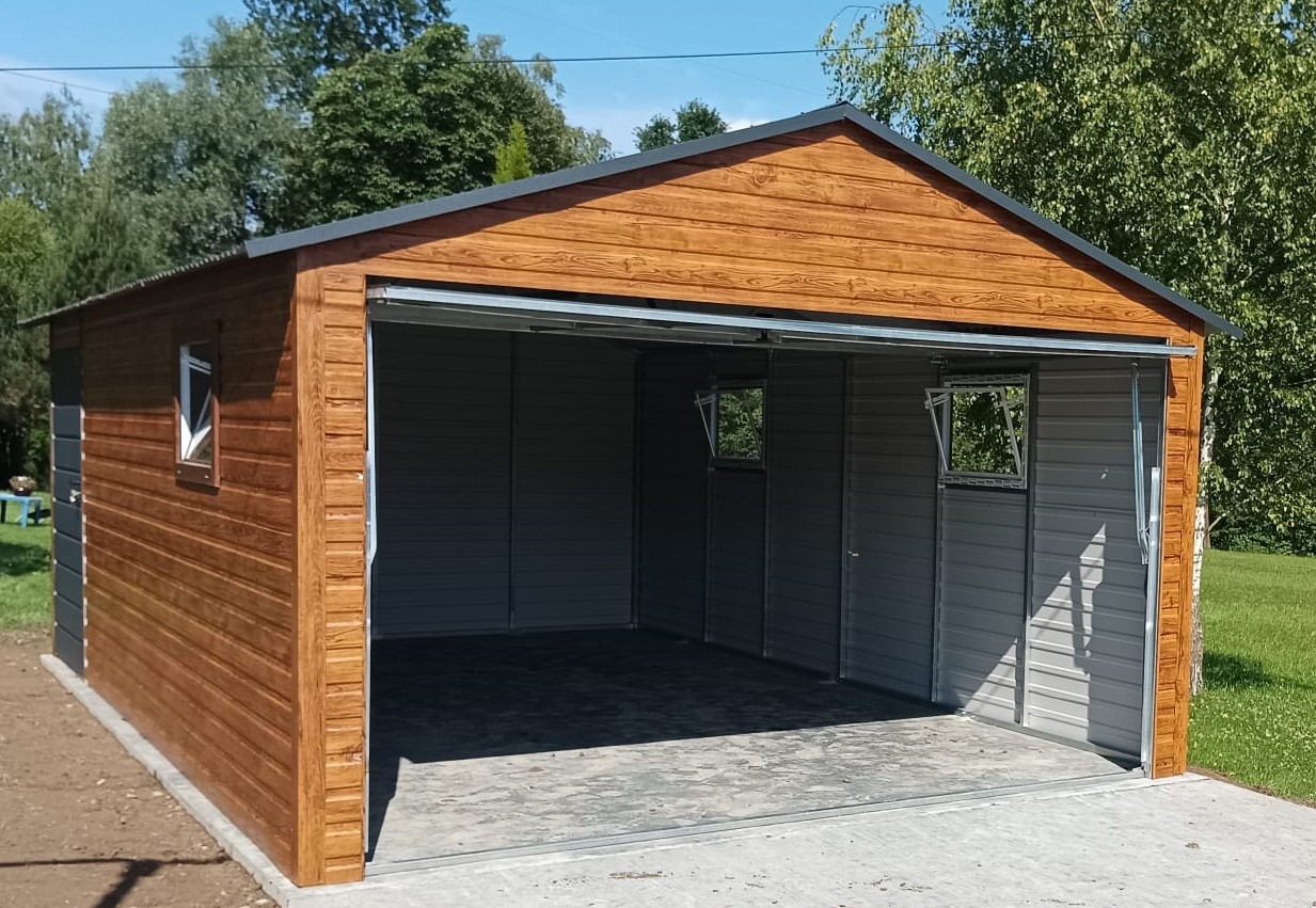 Zmontowany garaż blaszany z otwieraną bramą, obudowany drewnopodobną blachą z dwoma małymi oknami bocznymi, widok z zewnątrz na betonowy podjazd i zielone otoczenie.