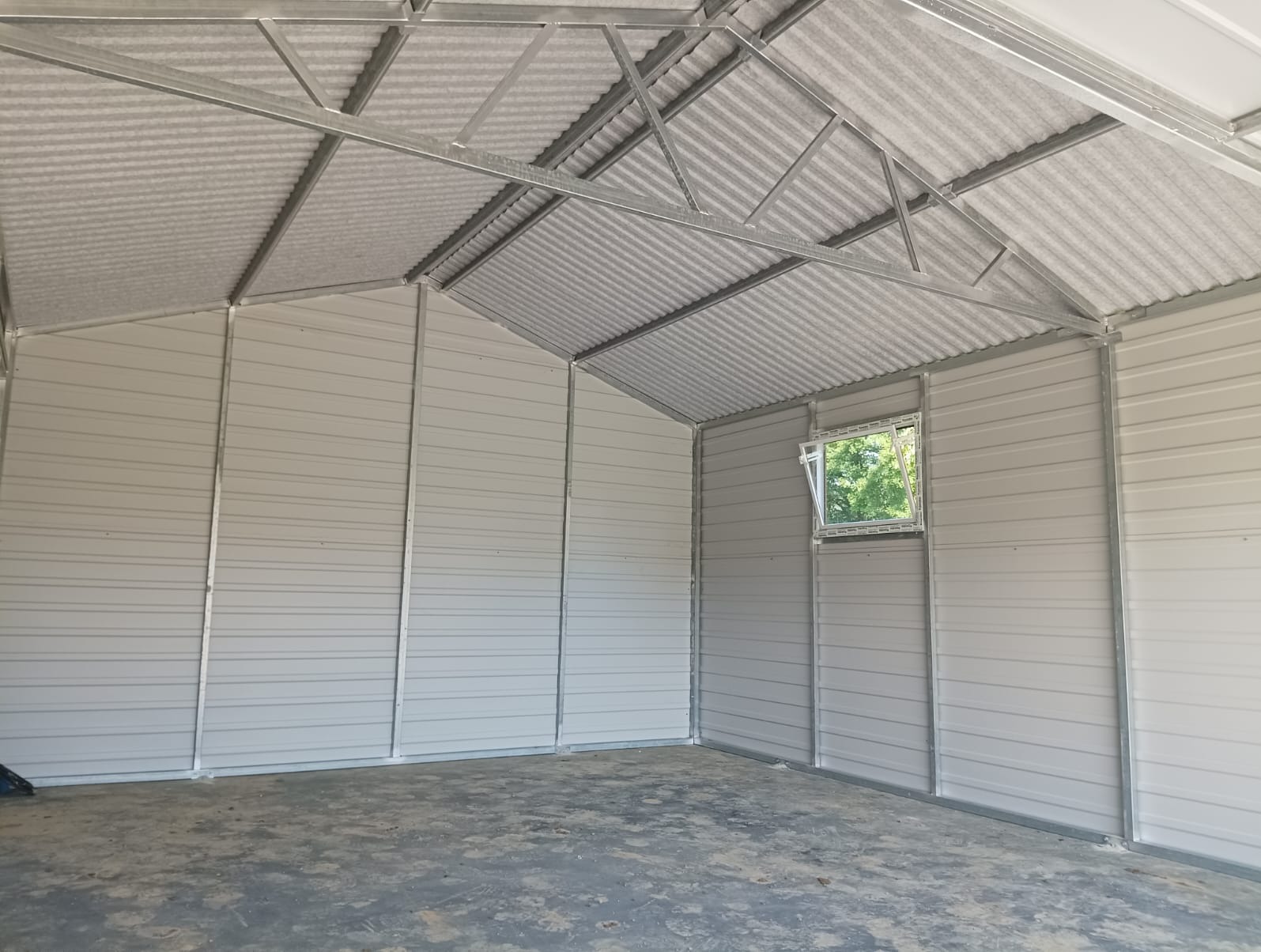 Wnętrze szarego, blaszanego garażu z otwartym oknem, ukazującym zieleń drzew na zewnątrz.
