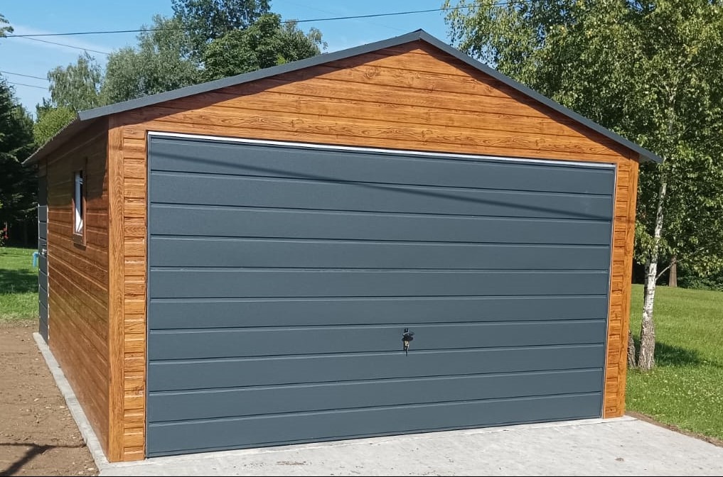 Gotowy garaż z drewnianą elewacją i szarą bramą segmentową, zamek na bramie, świeżo zagospodarowany teren wokół.