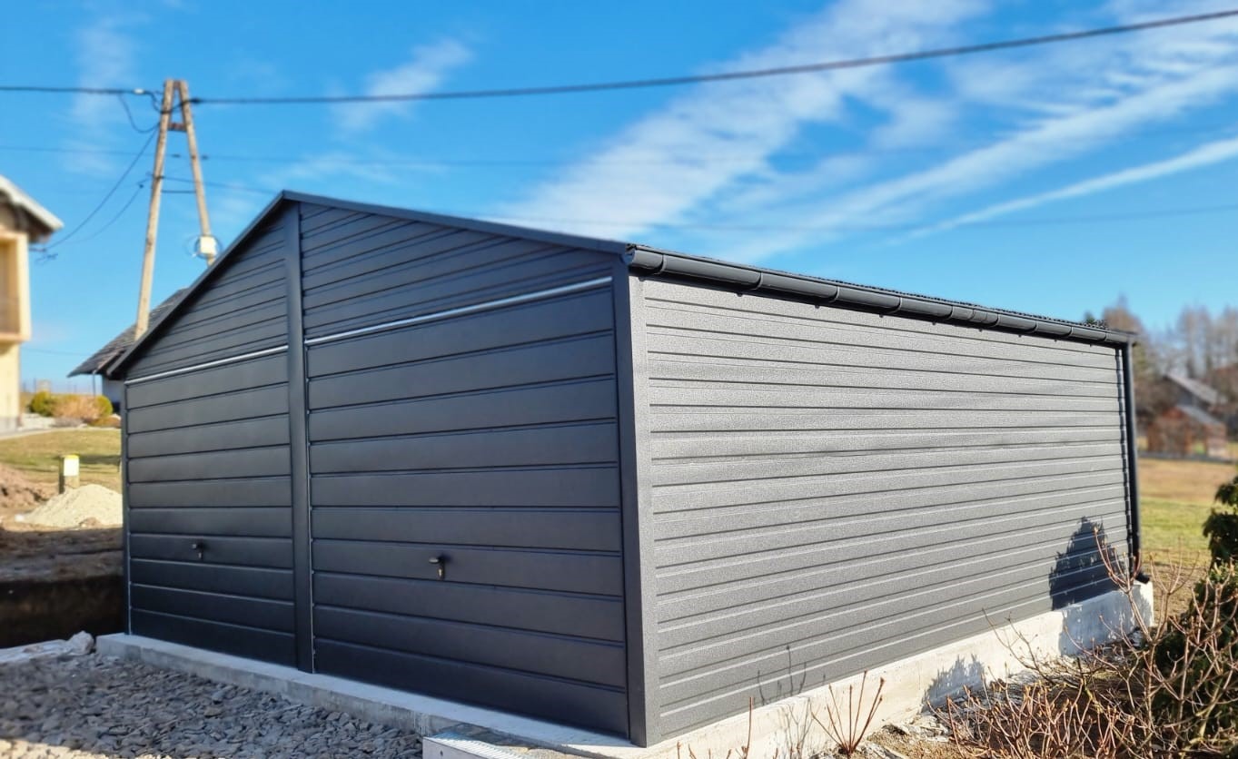 Ciemnoszary garaż blaszany z dwuspadowym dachem, posadowiony na betonowej podmurówce, widok z zewnątrz w słoneczny dzień.