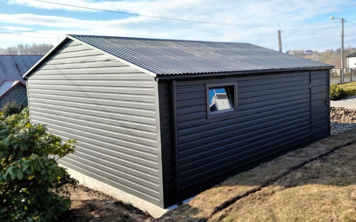 Nowoczesny, ciemnoszary garaż blaszany z oknem, wykończony poziomymi panelami, na betonowej podmurówce, z widocznym fragmentem dachu sąsiedniego budynku i błękitnym niebem w tle.