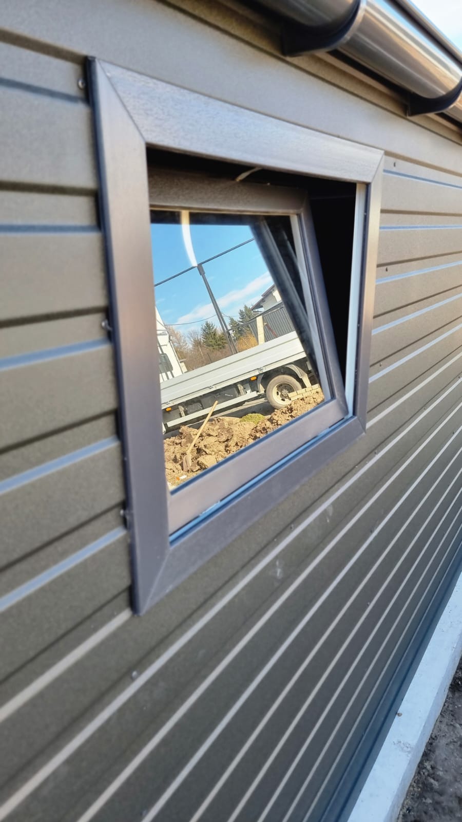 Szary garaż blaszany z uchylnym oknem, w którym odbija się krajobraz z zaparkowanym samochodem dostawczym i stertą ziemi ze szpadlem.