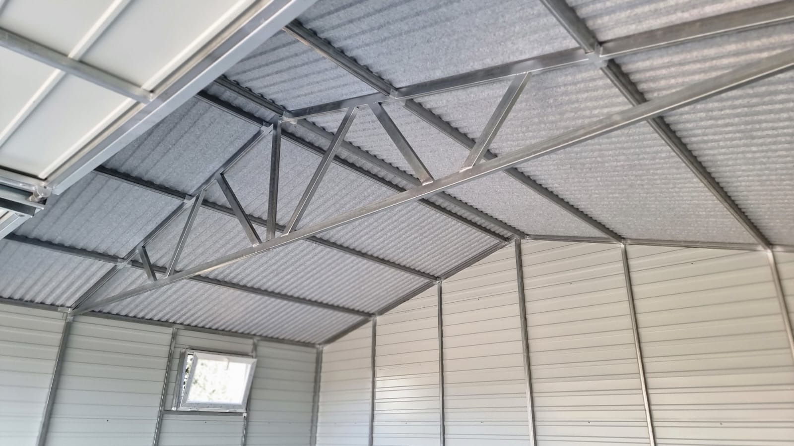 Wnętrze blaszanego garażu z widoczną konstrukcją dachu i ścianami z blachy falistej, okno w tylnej ścianie.
