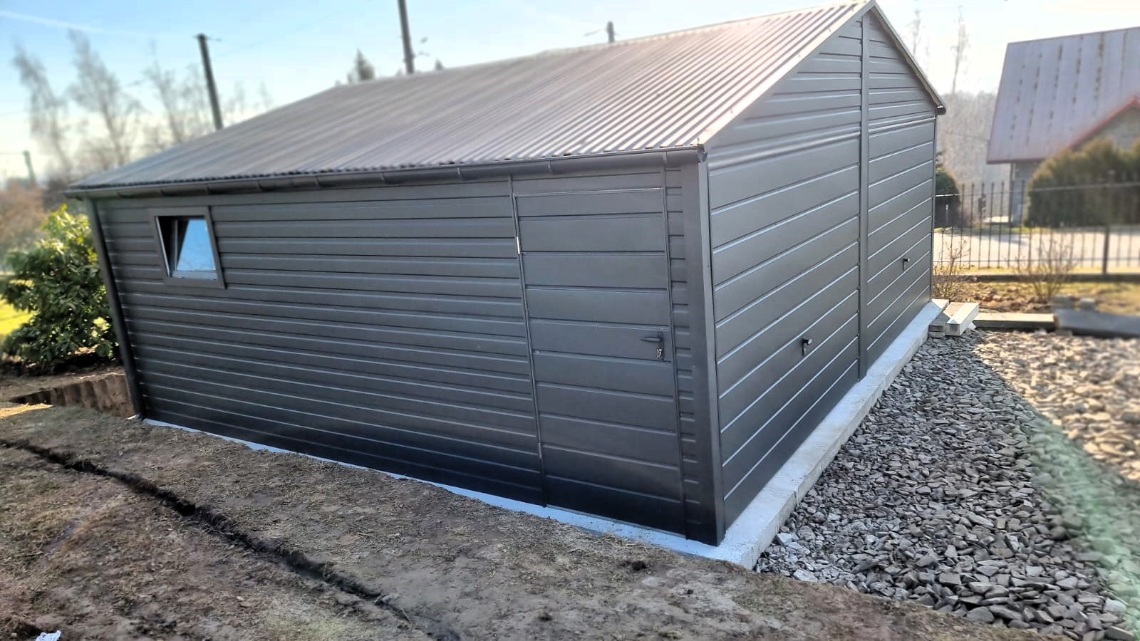 Nowoczesny, antracytowy garaż z blachy trapezowej, z małym oknem i drzwiami bocznymi, posadowiony na betonowej wylewce, z widoczną linią wykopu w ziemi i kamieniami ozdobnymi.