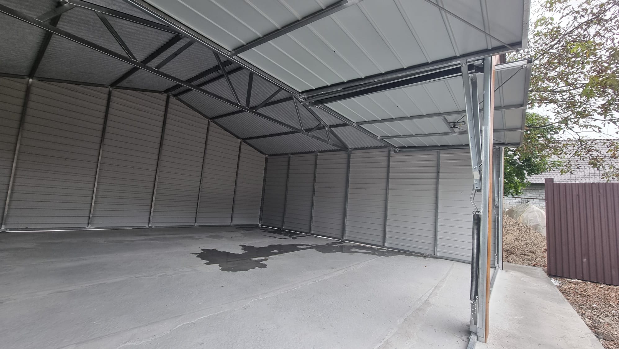 Wnętrze nowego, metalowego garażu z podnoszoną bramą, widoczne ślady wilgoci na betonowej posadzce.