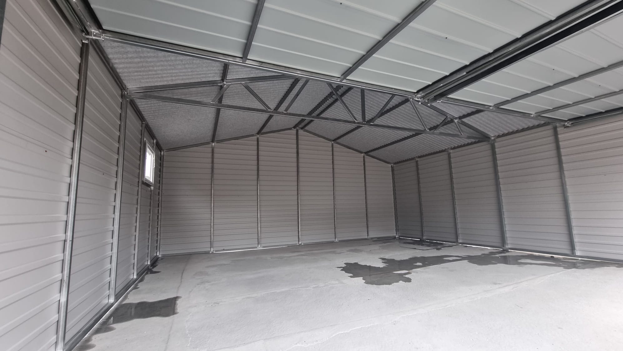 Wnętrze szkieletu metalowego garażu z widoczną konstrukcją dachu, ścianami z blachy trapezowej i betonową posadzką z ciemniejszymi plamami.