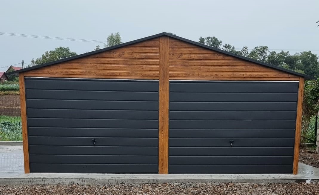 Dwustanowiskowy garaż blaszany z drewnianą górną częścią i grafitowymi, poziomymi panelami bram, widoczny na tle pola i pochmurnego nieba.