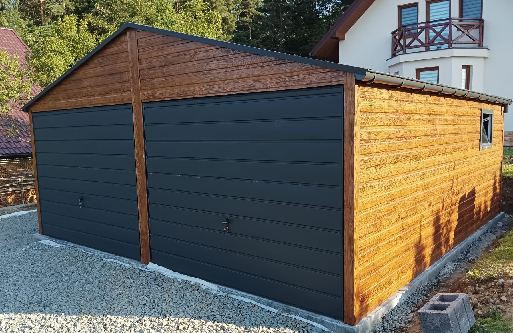 Dwustanowiskowy garaż blaszany z drewnopodobną elewacją i grafitowymi bramami segmentowymi, posadowiony na podsypce żwirowej, na tle domu jednorodzinnego.