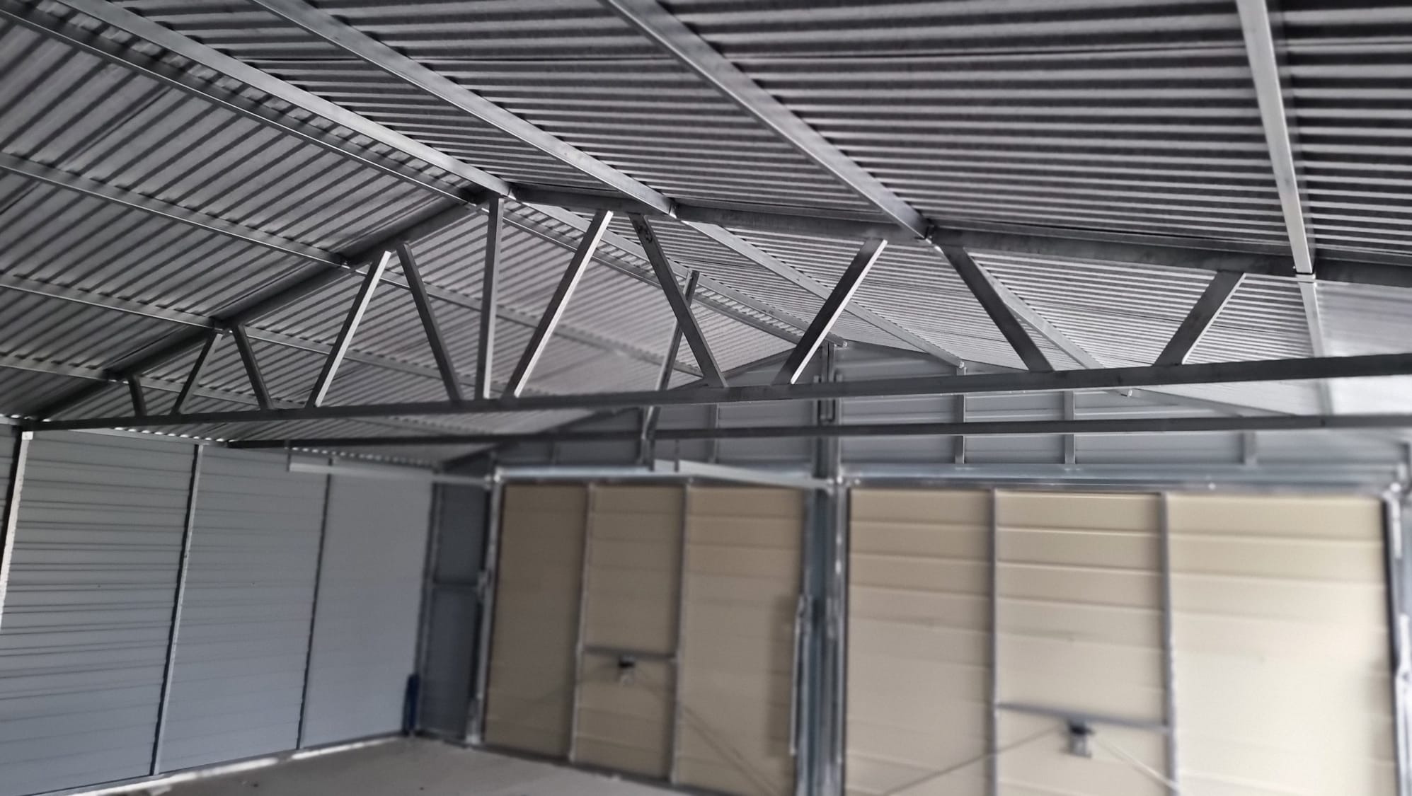 Wnętrze stalowej konstrukcji z widoczną blachą falistą dachu i metalowymi elementami nośnymi, ściany z paneli w odcieniach szarości i beżu.