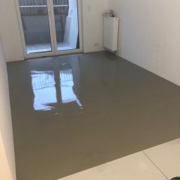 Tombud - Wylany, mokry samopoziomujący podkład podłogowy w jasnym pomieszczeniu z balkonem. Widoczny grzejnik i fragment białej podłogi z płytek.