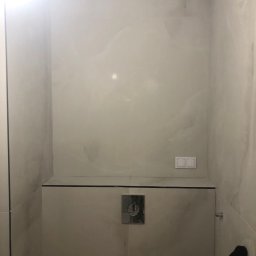 Tombud - Wykończona łazienka z beżowymi płytkami imitującymi marmur, widoczny panel podtynkowy WC i włącznik światła. Górna krawędź ściany zabezpieczona niebieską taśmą.