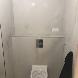 Tombud - Wykończona toaleta podwieszana w małej łazience, obłożona jasnymi płytkami imitującymi marmur, z czarnym akcentem półki i wiszącym czarnym szlafrokiem.