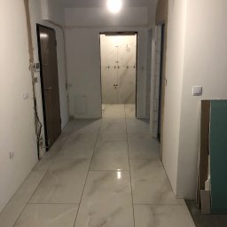Tombud - Korytarz w trakcie wykończenia w Zamościu: ułożone płytki podłogowe, przygotowane ściany do malowania, widoczne instalacje elektryczne i sufit podwieszany.