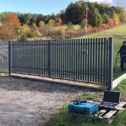 Tombud - Montaż nowoczesnego, metalowego ogrodzenia panelowego z bramą wjazdową na tle jesiennego krajobrazu. Widoczne narzędzia i ekipa montażowa.