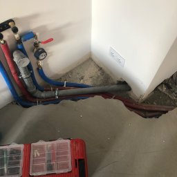 Tombud - Instalacja wodno-kanalizacyjna w rogu pomieszczenia, widoczne rury w kolorach niebieskim i czerwonym, skrzynka narzędziowa, gniazdko elektryczne na ścianie.