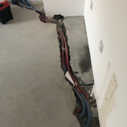 Tombud - Wykop w szarej posadzce z widocznymi rurami instalacji wodnej i grzewczej, częściowo zakrytymi. Na ścianie widoczne puszki elektryczne i włączniki.