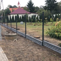 Tombud - Nowoczesne ogrodzenie panelowe z podmurówką w kolorze grafitowym, oddzielające posesję od chodnika i drogi. W tle widoczny dom z czerwonym dachem i zielenią.