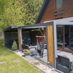 Tombud - Nowoczesna pergola z szarymi słupami i drewnianymi panelami, wyposażona w wiszący hamak i meble ogrodowe, przylegająca do drewnianego domu z zielonym trawnikiem.