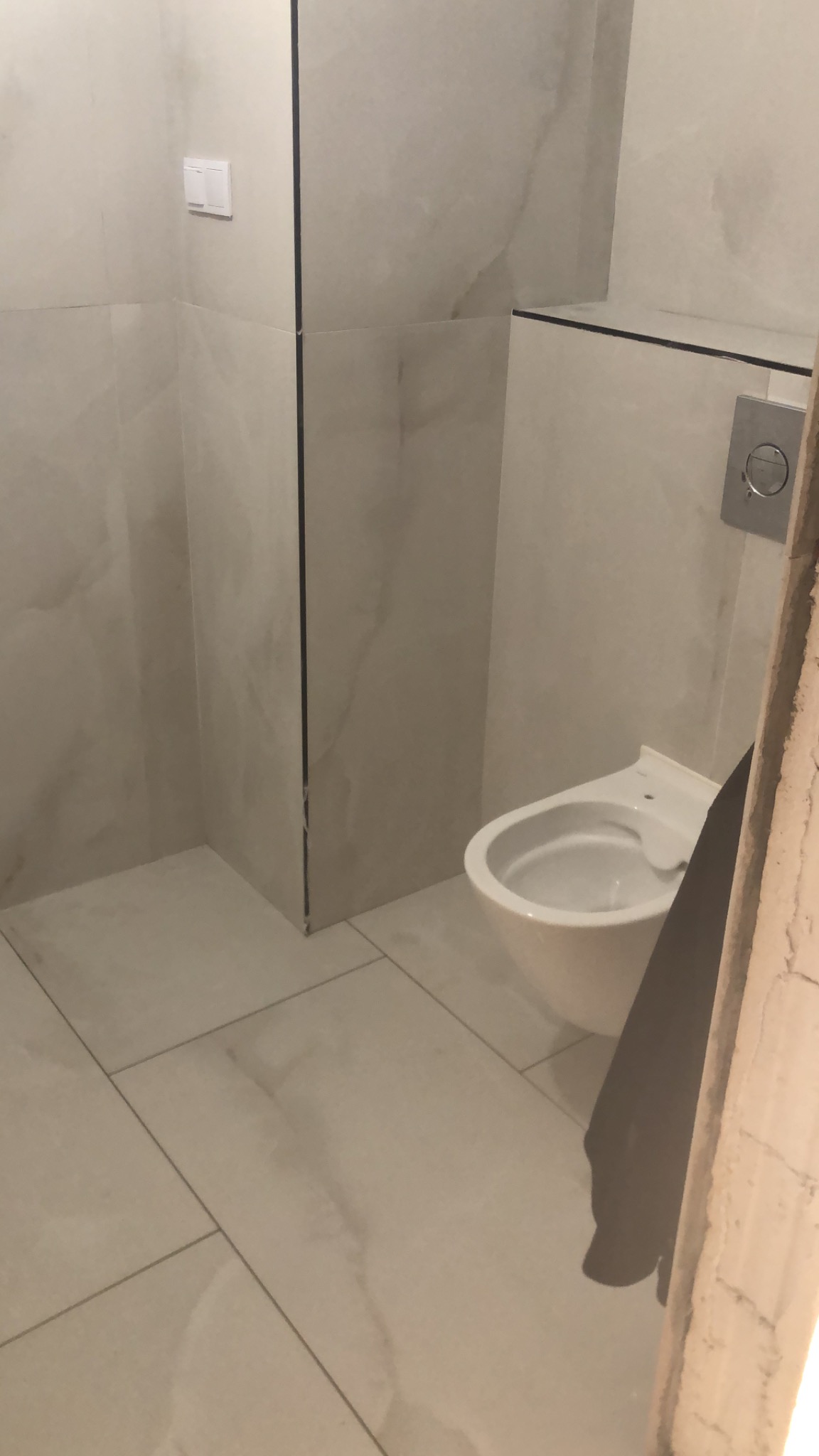 Wykończona łazienka z podwieszaną toaletą i płytkami imitującymi marmur. Widoczny fragment futryny drzwi. Minimalistyczny design.