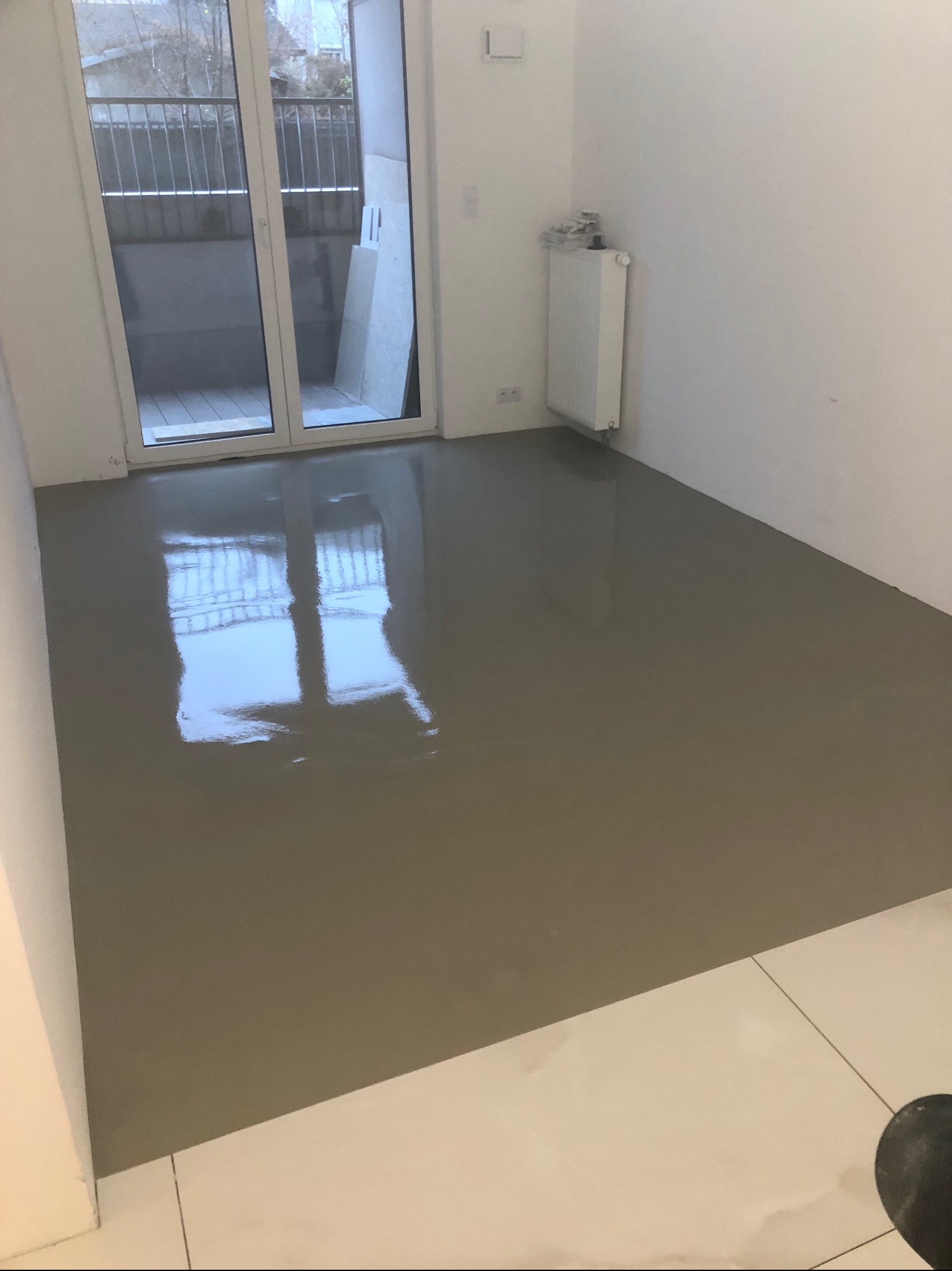 Wylany, mokry samopoziomujący podkład podłogowy w jasnym pomieszczeniu z balkonem. Widoczny grzejnik i fragment białej podłogi z płytek.