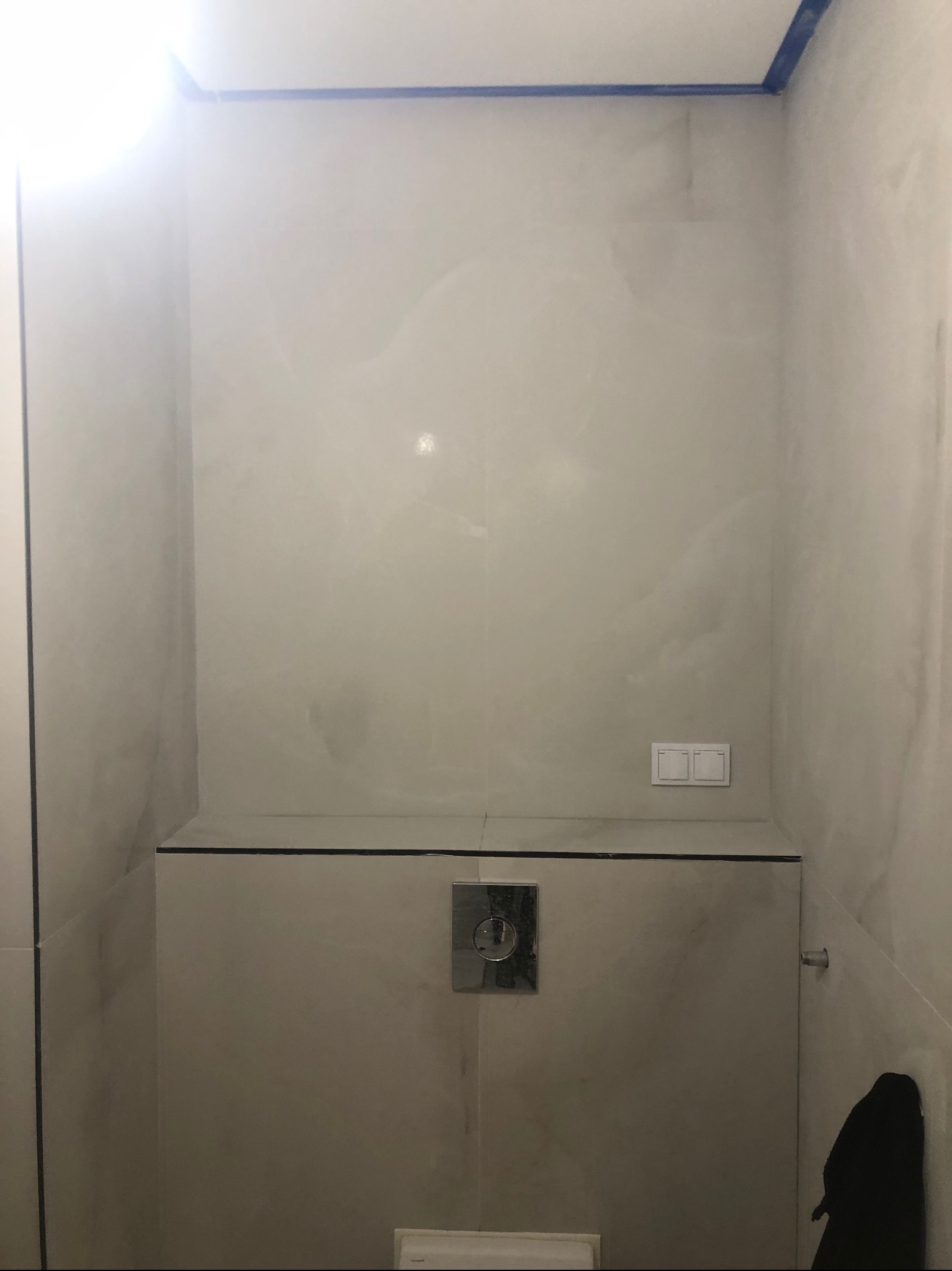 Wykończona łazienka z beżowymi płytkami imitującymi marmur, widoczny panel podtynkowy WC i włącznik światła. Górna krawędź ściany zabezpieczona niebieską taśmą.