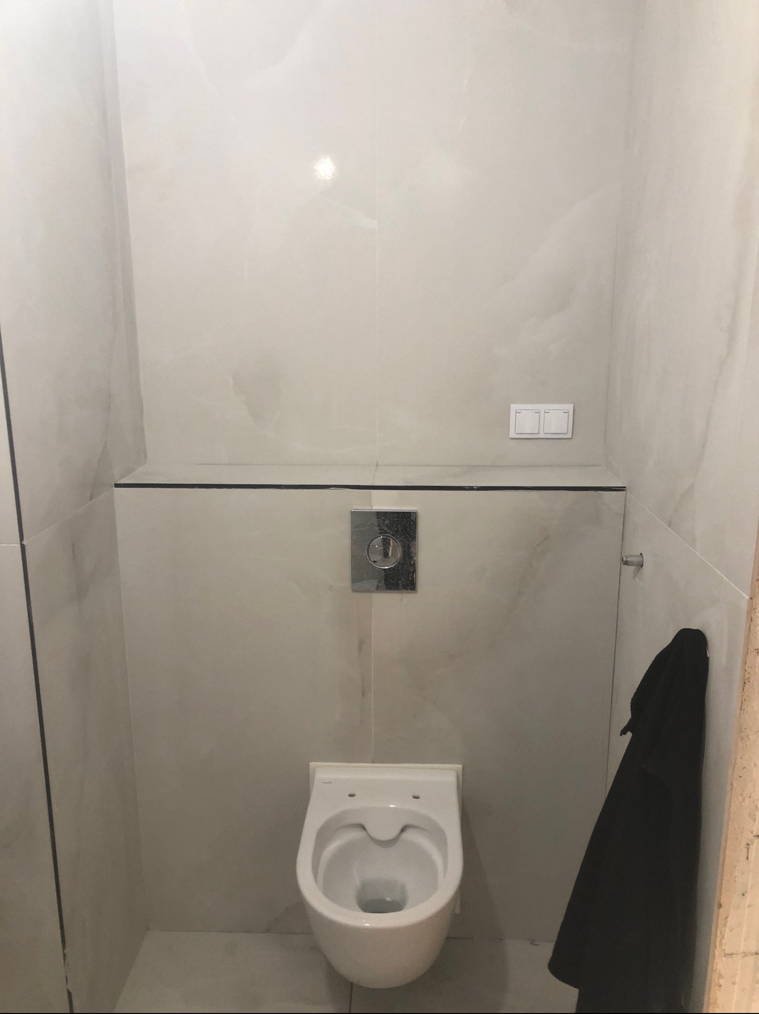 Wykończona toaleta podwieszana w małej łazience, obłożona jasnymi płytkami imitującymi marmur, z czarnym akcentem półki i wiszącym czarnym szlafrokiem.