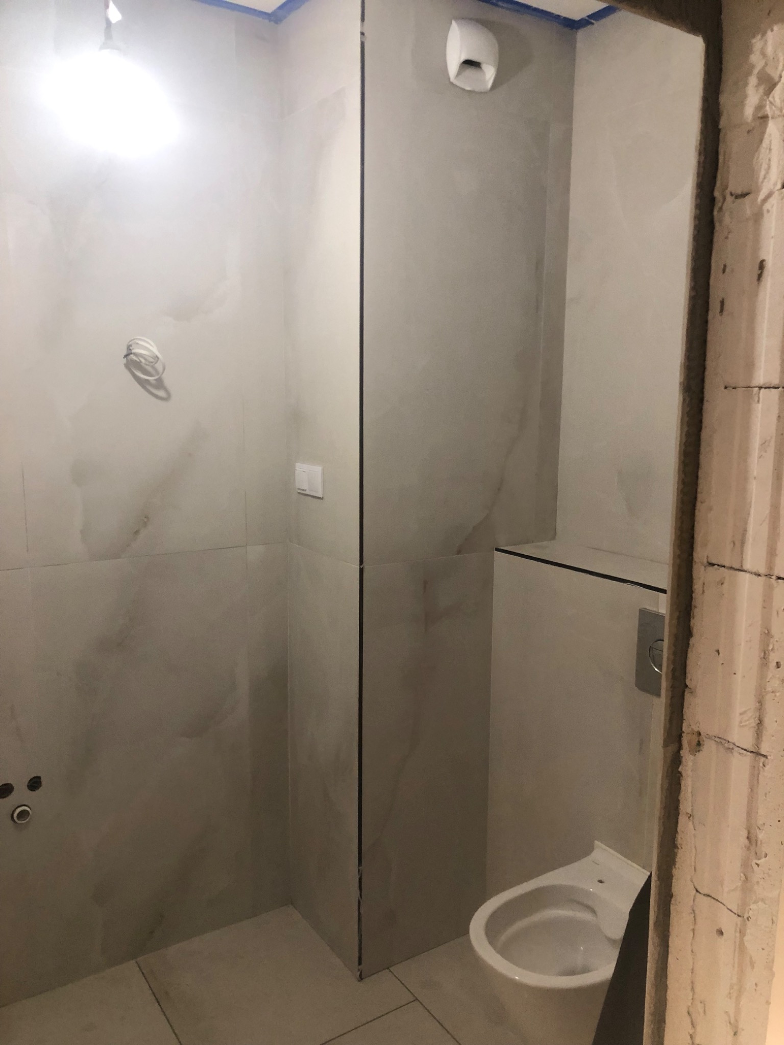 Wnętrze łazienki w trakcie wykończenia: szare płytki imitujące marmur na ścianach, częściowo zamontowana toaleta podwieszana, widoczne instalacje i surowy otwór drzwiowy.