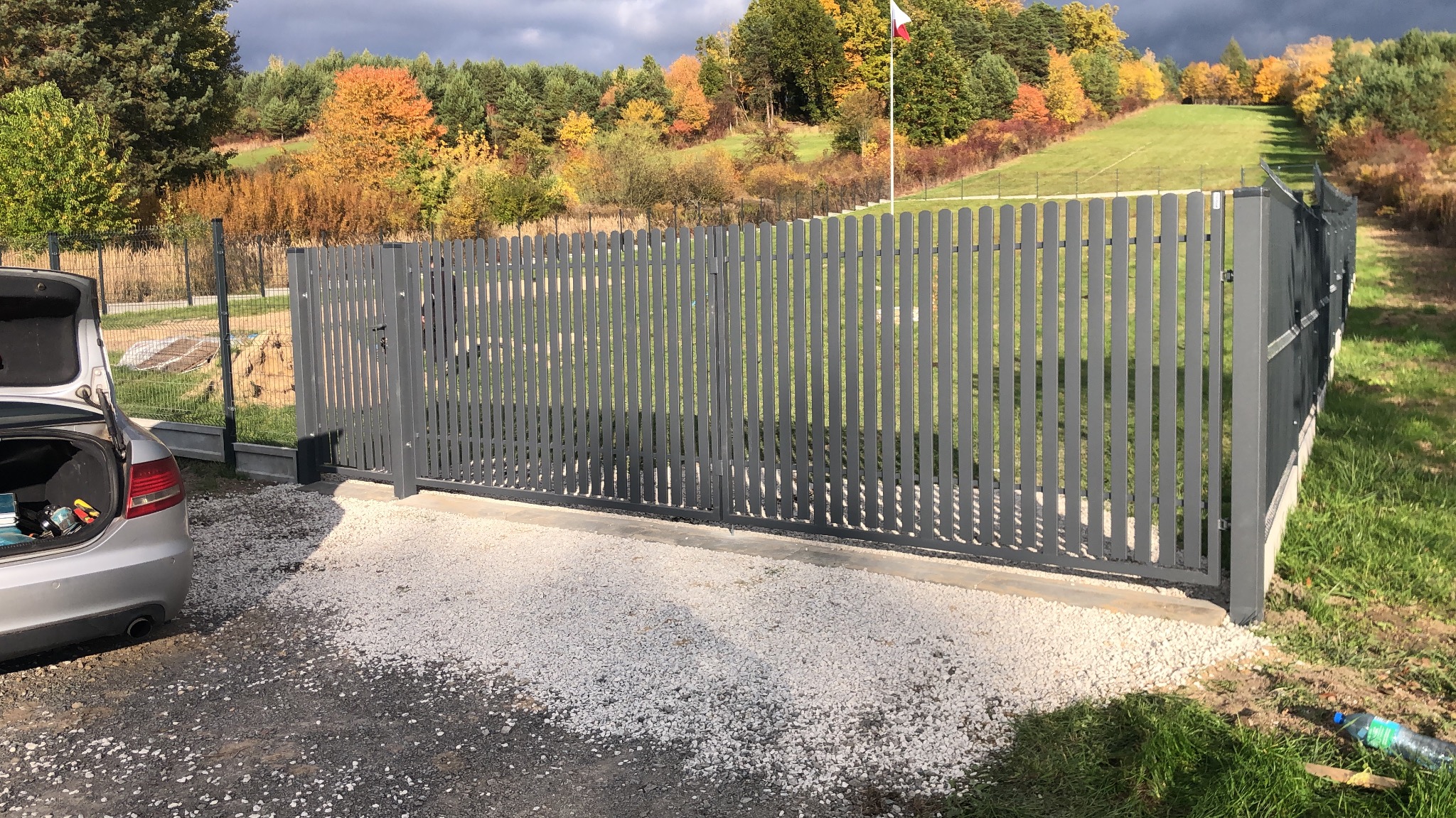Nowoczesna, szara brama wjazdowa z pionowymi szczeblami, świeżo zamontowana na posesji z widokiem na jesienny krajobraz i polską flagę w tle.
