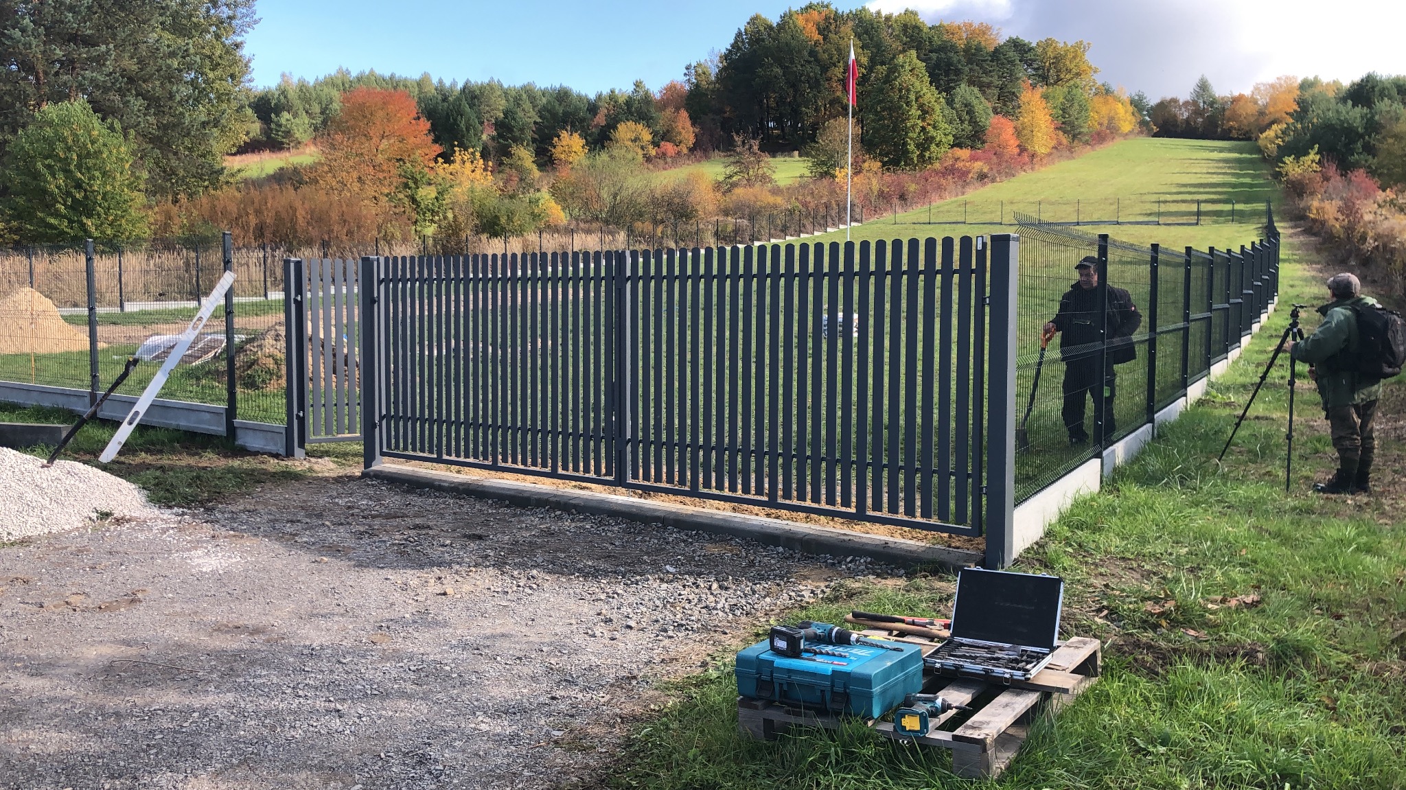Montaż nowoczesnego, metalowego ogrodzenia panelowego z bramą wjazdową na tle jesiennego krajobrazu. Widoczne narzędzia i ekipa montażowa.