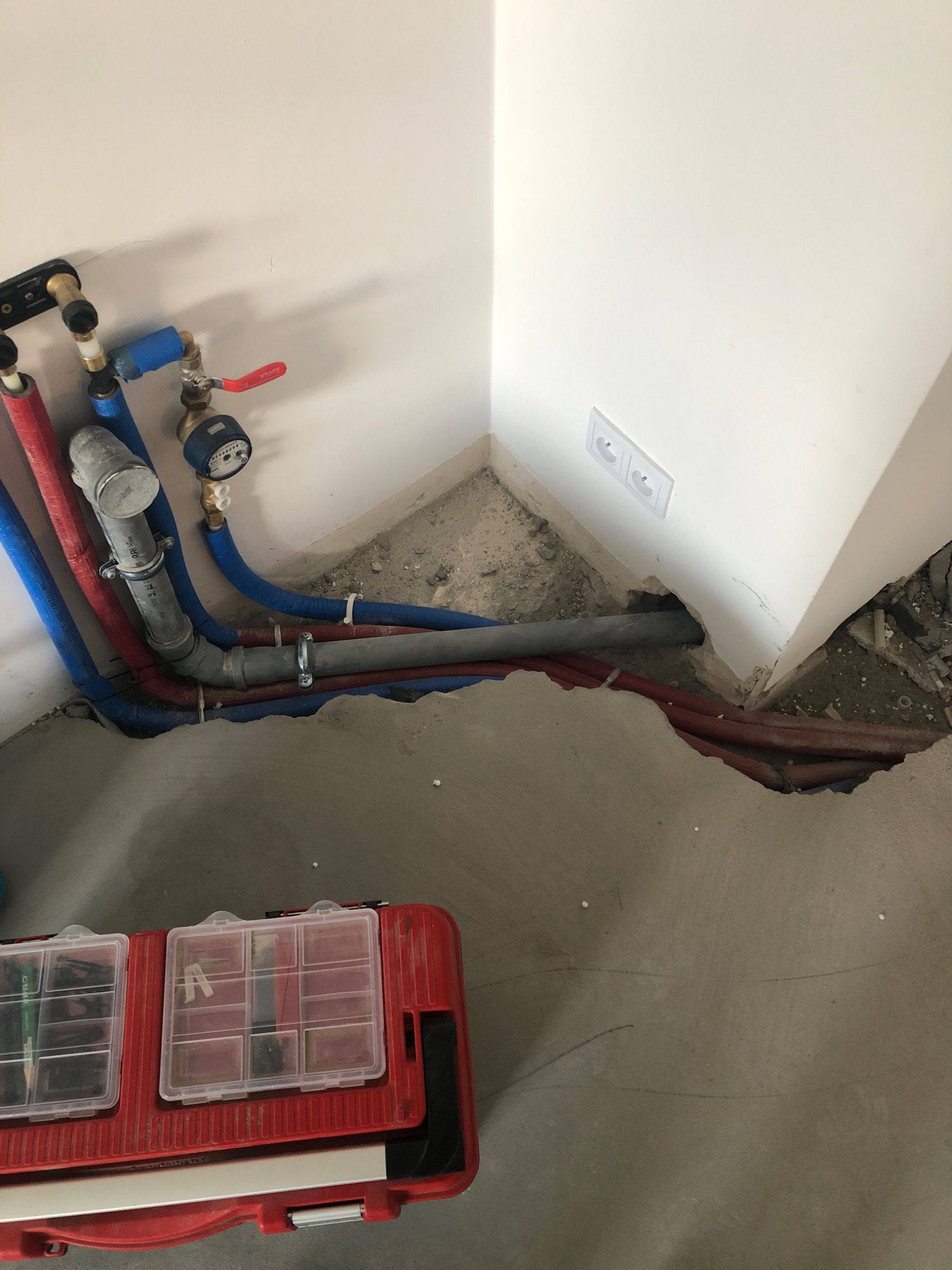 Instalacja wodno-kanalizacyjna w rogu pomieszczenia, widoczne rury w kolorach niebieskim i czerwonym, skrzynka narzędziowa, gniazdko elektryczne na ścianie.